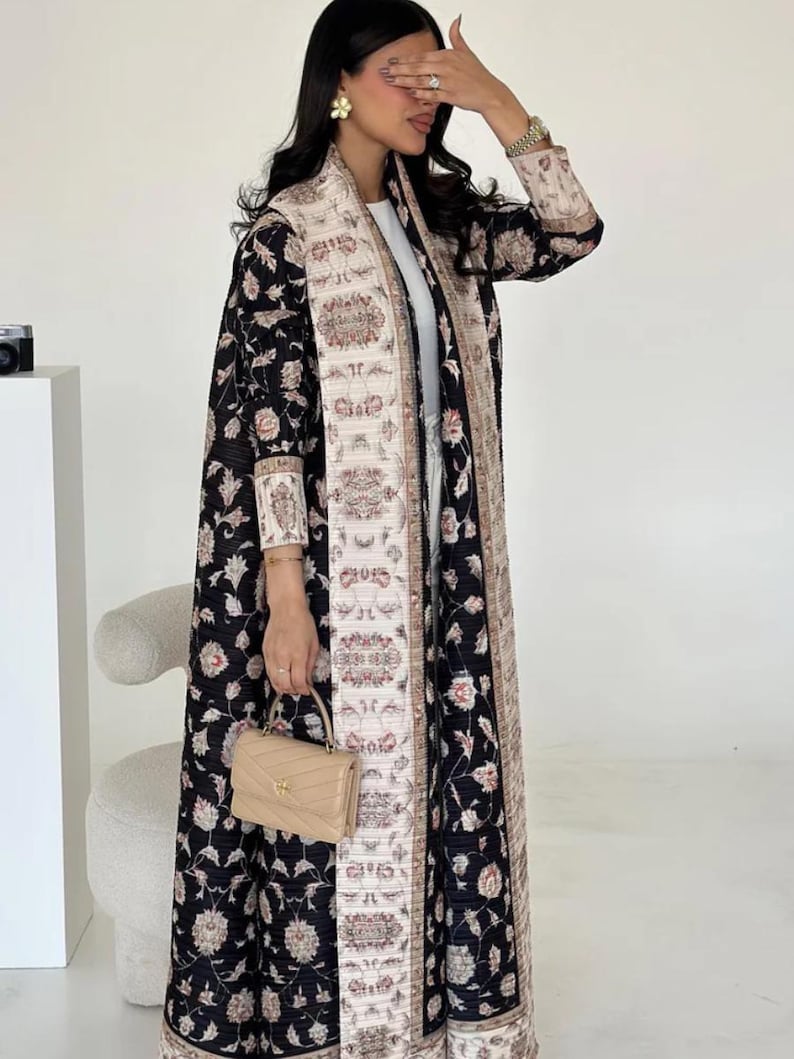 K&ouml;nnte beinhalten: Ein langer, schwarzer Mantel mit floralem Muster und einem cremefarbenen, gemusterten Rand und Futter. Der Mantel wird &uuml;ber einem wei&szlig;en Outfit getragen und mit einer kleinen, beigen Handtasche kombiniert. Das Blumenmuster enth&auml;lt rosa und cremefarbene Blumen.