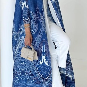 May include: A long, flowing blue and white paisley patterned coat. The coat is worn over white trousers and a white top. A small beige handbag is held in the hand. The coat is a statement piece.