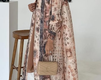May include: A long, patterned coat in shades of pink, brown, and blue with a floral and abstract design. The coat is open in the front and has long sleeves. A small, tan handbag is held in the hand.