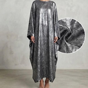 May include: A floor-length, loose-fitting kaftan dress in a metallic silver color with a crinkled, textured finish. The garment features long, wide sleeves and a round neckline, with an inset circular detail showing the fabric texture.