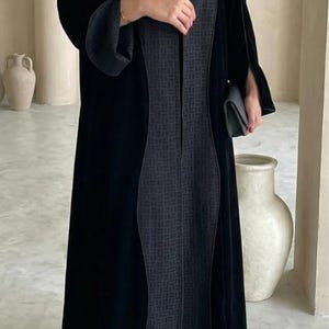 May include: A long black velvet abaya with a dark gray patterned panel down the front. The abaya has long sleeves and is paired with a small black clutch and embellished shoes. The background features neutral-toned walls and decorative vases.