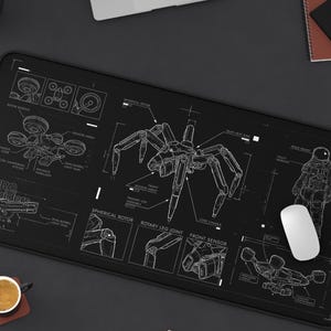 Könnte beinhalten: Schwarzes rechteckiges Mauspad mit einem weißen schematischen Design eines spinnenartigen Roboters, eines Astronauten und anderer mechanischer Komponenten. Eine weiße Maus liegt auf dem Pad. Ein Laptop, eine Kaffeetasse und Bleistifte sind ebenfalls sichtbar.