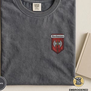 Puede incluir: Camiseta de color grafito con cuello redondo. La camiseta presenta un parche bordado con la palabra "Barbarian" encima de un diseño de escudo rojo con hachas cruzadas. Fabricada en material Comfort Colors.
