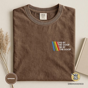 以下が含まれることがあります： エスプレッソ色のTシャツ。クルーネックと半袖。カラフルな刺繍デザインで「LIVE BY THE GOOP DIE BY THE GOOP」の文字が施されています。柔らかく快適な素材で作られています。