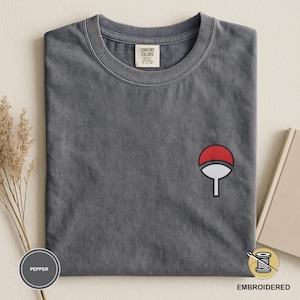 May include: A gray, short-sleeved t-shirt with a small, embroidered red and white emblem on the chest. The shirt has a crew neck and a tag that reads "Comfort Colors". The word "Embroidered" is printed on the bottom right.