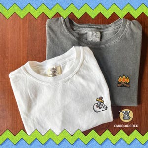 Pode incluir: Duas camisetas, uma branca e outra cinza, são exibidas. A camiseta branca apresenta uma caveira pixelizada bordada com uma espada, enquanto a camiseta cinza tem um design de fogueira pixelizada. Ambas as camisetas têm a etiqueta "Comfort Colors".