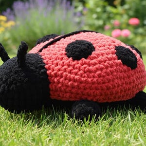 May include: A handmade, crocheted ladybug toy. The ladybug is red with black spots and a black head and legs. It is sitting on a bed of green grass, with a blurred background of flowers and foliage.