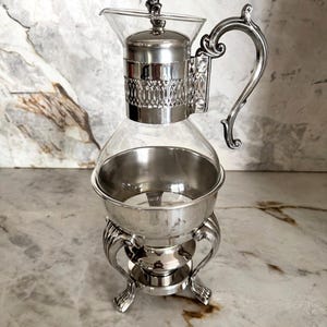 Puede incluir: Una cafetera plateada con una jarra de cristal y un asa ornamentada. La cafetera tiene una base decorativa y una tapa con un pequeño adorno. El diseño es vintage y elegante, adecuado para servir café o té.