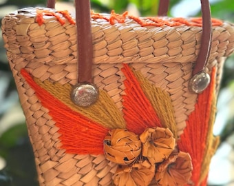 Könnte beinhalten: Gewebte Strohhandtasche mit braunen Ledergriffen und Akzenten. Die Tasche hat orange und gelbe Stickereien, darunter ein Sonnenstrahl-Design und dekorative Blumenakzente. Silberfarbene Metallnieten sichern die Griffe.