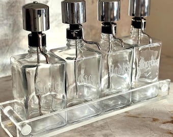 Op de afbeelding: Een set van vier heldere glazen drankflessen met zilverkleurige doppen en schenktuiten, gehouden in een helder acrylrek. Elke fles is gelabeld met een ander dranktype.