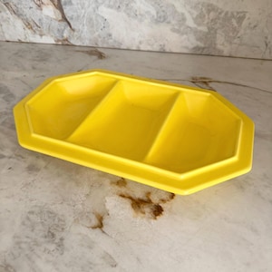 May include: A bright yellow serving tray with an octagonal shape and three distinct rectangular compartments. The tray features a wide, flat rim and angled interior walls, providing a structured design for organizing small items or food portions.