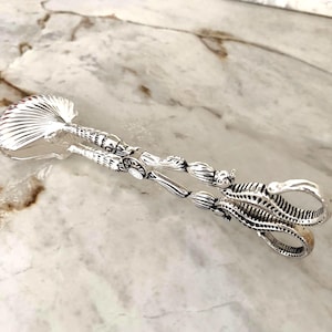 May include: Silver-toned serving tongs with intricate seashell and sea creature designs. The tongs feature a shell-shaped scoop and a detailed handle with an octopus-like grip. The design is ornate and elegant.