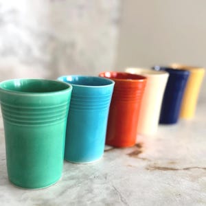 Könnte beinhalten: Reihe von sechs bunten Keramikbechern in Grün, Blau, Rot, Creme, Marineblau und Gelb. Jeder Becher hat ein geriffeltes Design am oberen Rand und eine glatte, zylindrische Form. Die Becher sind in einer Reihe angeordnet und zeigen ihre leuchtenden Farben.