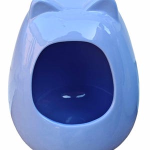 May include: A light blue ceramic pet bed shaped like an egg with cat ears on top. The opening is a dark blue color. The bed has a glossy finish and is designed for small pets.