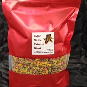 Puede incluir: Una bolsa roja de comida para petauros del azúcar con una ventana transparente que muestra la colorida mezcla de comida y golosinas. La etiqueta dice "Sugar Glider Extreme Blend" y "1 Pound Bag".