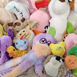 May include: A selection of colourful plush dog toys in various shapes and sizes. Includes a yellow pineapple, a purple octopus, a blue whale, and a white dog-shaped toy. Some toys have text like "My M".