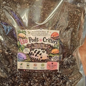 Puede incluir: Una bolsa de plástico transparente que contiene una mezcla de tierra marrón oscuro para isópodos, con una etiqueta que muestra el texto Iso Pods-N-Critters, Isopod Soil Prepared by Hand y una lista de ingredientes como musgo y madera.