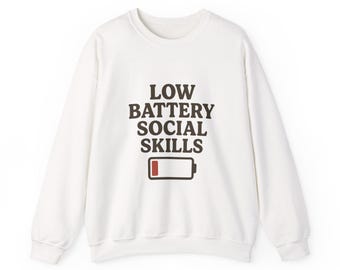 Puede incluir: Sudadera blanca con el texto "LOW BATTERY SOCIAL SKILLS" en letras marrones oscuras. Debajo, un gráfico de batería baja. La sudadera tiene mangas largas y cuello redondo.