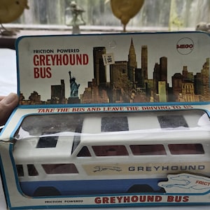 May include: A vintage friction-powered Greyhound bus toy inside its original cardboard packaging. The box features a graphic of the New York City skyline, the Statue of Liberty, and the text "Friction Powered Greyhound Bus" and "Take the bus and leave the driving to us."