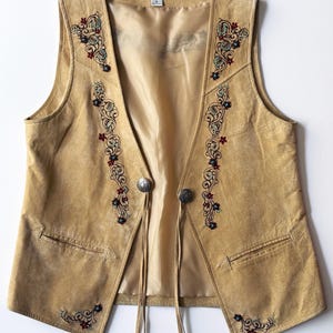 May include: A tan suede vest featuring intricate floral embroidery in red, blue, and black along the front panels. The garment includes two silver-toned metal buttons at the center with long suede fringe ties and a smooth tan interior lining.