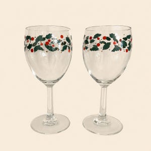 May include: Two clear glass wine glasses featuring a painted band around the upper bowl. The design consists of dark green holly leaves and bright red berries arranged in a continuous horizontal pattern encircling the glass stemware.