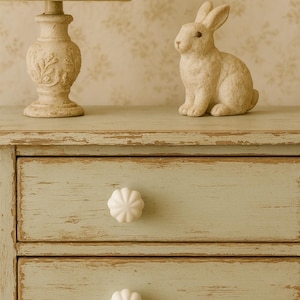 Könnte beinhalten: Eine hellgrüne, abgenutzte Holzkommode mit zwei Schubladen, jede mit einem weißen, blütenförmigen Knauf. Eine cremefarbene Lampe mit floralem Lampenschirm und eine kleine Hasenfigur stehen obenauf. Der Hintergrund ist eine Blumentapete.
