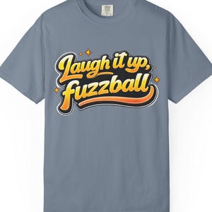 May include: A dusty blue t-shirt with the phrase "Laugh it up, Fuzzball" in a retro, layered design. The text is in yellow, orange, and black, with white outlines and star accents. The shirt is a casual, short-sleeved style.