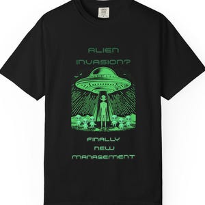 May include: Black t-shirt with a green graphic of a UFO, an alien, and smaller alien figures. The text "ALIEN INVASION? FINALLY NEW MANAGEMENT" is printed in green. The shirt has short sleeves and a crew neck.