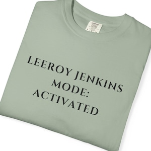 Może przedstawiać: Szałwiowa zielona koszulka z czarnym napisem "LEEROY JENKINS MODE: ACTIVATED". Koszulka jest złożona i ma metkę w prawym górnym rogu. Koszulka jest wykonana z miękkiego materiału.