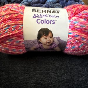 May include: A large ball of Bernat Softee Baby Colors yarn in shades of pink, red, yellow, and blue. The yarn is wrapped around a label featuring a baby. The yarn is on a dark blue surface.