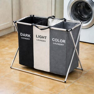 Może przedstawiać: Trzysekcyjny kosz na pranie z metalową ramą w kolorze srebrnym. Każda sekcja oznaczona jest napisami "DARK LAUNDRY", "LIGHT LAUNDRY" i "COLOR LAUNDRY". Kosz posiada czarne, białe i szare przegrody. W tle widoczna jest pralka.
