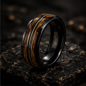 May include: A black tungsten ring featuring a central inlay of brown wood grain and a thin gold-colored guitar string detail. The interior of the band is clearly engraved with the word TUNGSTEN in a rectangular border, showcasing a polished finish.