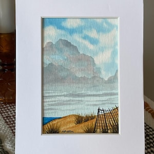 May include: Watercolor painting of a coastal landscape, framed in white. The artwork depicts a beach scene with a cloudy sky, ocean, and a fence. The painting is signed "MP Patton".