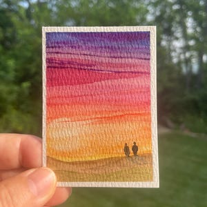 May include: A small rectangular watercolor painting on textured paper featuring a vibrant sunset gradient transitioning from deep purple at the top to pink, orange, and yellow, with two small dark silhouettes standing on a light brown landscape.