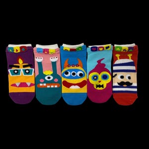 May include: A set of five ankle socks featuring colorful monster faces. The socks are purple, pink, blue, light blue, and red, each with unique eyes, mouths, and decorative heart-patterned cuffs at the top. The designs include various expressions.
