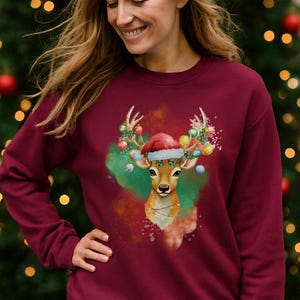 Op de afbeelding: Bordeauxkleurige crewneck sweatshirt met een illustratie van een hert met een kerstmuts en feestelijke versieringen op zijn gewei. Het hert staat tegen een groene en rode aquarel achtergrond.