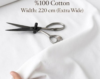 May include: A roll of white cotton fabric with the text "%100 Cotton Width: 220 cm (Extra Wide)". A pair of black and silver scissors rests on the fabric. A hand holds a piece of the fabric.