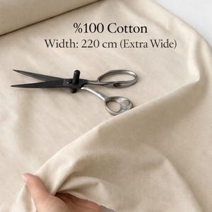 May include: A roll of cream-coloured cotton fabric with the text "%100 Cotton" and "Width: 220 cm (Extra Wide)". A pair of black and silver scissors lies on the fabric. A hand holds the fabric.