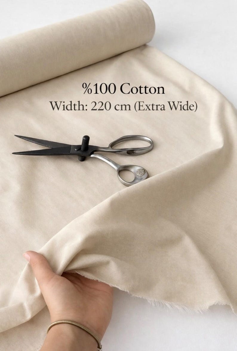 May include: A roll of beige cotton fabric with the text "%100 Cotton" and "Width: 220 cm (Extra Wide)". A pair of black and silver scissors rests on the fabric. A hand holds the fabric.
