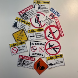 May include: A collection of safety signs with various warnings and instructions. The signs include "Danger," "Caution," and "Notice" labels, with symbols indicating required safety equipment and prohibited activities. The signs are in various shapes and sizes.