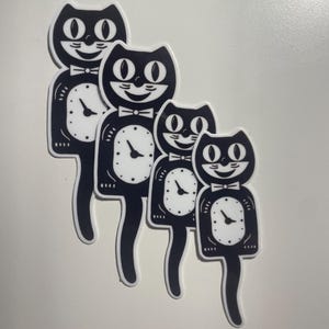 May include: Four black and white cat-shaped stickers. Each cat has a clock face for a body, a bow tie, and a smiling face. The stickers are arranged diagonally on a white background.