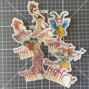 May include: A collection of die-cut stickers featuring illustrations of a girl with red hair in various fancy outfits. Each sticker has the words "Fancy Nancy" printed on it.