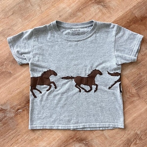 May include: A gray short-sleeved t-shirt featuring a brown horse graphic. The shirt has a crew neck and is made of a soft material. The horse graphic is repeated across the front of the shirt.