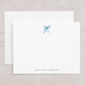 May include: White stationery set featuring a light blue watercolor bow design. The set includes a folded card and an envelope. The name "AMANDA DRAKE" is printed in a simple font at the bottom of the card.