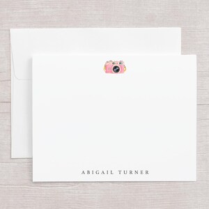 May include: White stationery set featuring a pink camera illustration. The set includes a blank card with the name "ABIGAIL TURNER" printed at the bottom, and a matching envelope. The camera illustration has yellow and pink accents.