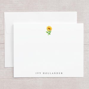 May include: White stationery set featuring a watercolor sunflower design. The set includes a blank card with a sunflower illustration and the text "IVY HOLLANDER", along with a matching envelope. The stationery is ideal for sending personalized notes.