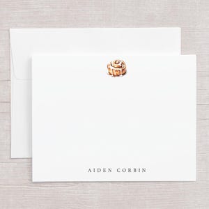 May include: White stationery set featuring a watercolor illustration of a cinnamon roll. The set includes a folded card and an envelope. The name "AIDEN CORBIN" is printed in a simple font at the bottom of the card.
