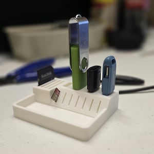 May include: A white plastic organiser with slots and a tray, holding a green and silver USB drive, a black USB charger, and a blue USB drive. A SanDisk SD card is also in a slot.