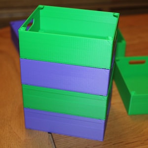May include: A stack of green and purple storage boxes with handle cutouts. One box is separate, next to the stack. The boxes are made of a solid material and are on a wooden surface. The boxes are empty.