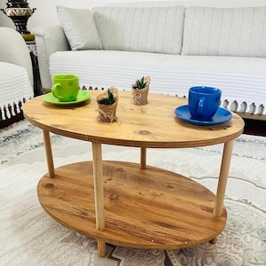 May include: Oval wooden coffee table with a lower shelf, supported by four cylindrical legs. The table has a natural wood finish and holds a green teacup and saucer, a blue teacup and saucer, and small potted plants.